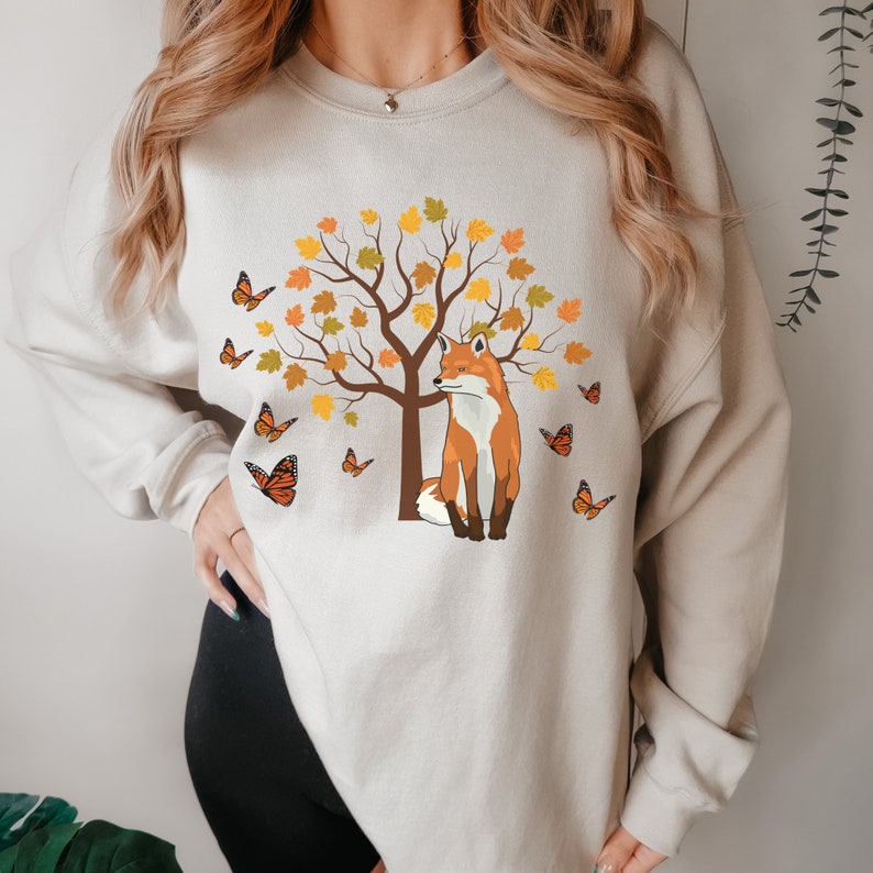 May include: A beige sweatshirt with a graphic of a fox sitting under a tree with orange and yellow leaves. There are butterflies flying around the tree.