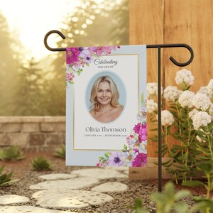 Può includere: Una bandiera da giardino con un ritratto di donna, circondata da composizioni floreali rosa, viola e bianche. La bandiera include il testo "Celebrating the Life of Olivia Thomson" e le date di nascita e di morte. Esposta su un supporto in metallo nero.