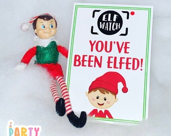 Può includere: Un biglietto bianco con bordi verdi e il testo "Elf Watch You've Been Elfed!" Un elfo dei cartoni animati con un cappello rosso è raffigurato sulla carta. Un elfo giocattolo con un cappello rosso e pantaloni a righe è seduto su una superficie bianca e soffice.