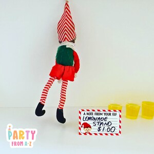 May include: A red and white striped elf doll hanging upside down from a ceiling. A sign reads "A note from your elf Lemonade Stand £1.00". Three yellow plastic cups are on a white surface.