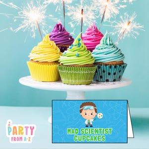 May include: A display of six cupcakes with colorful frosting, including yellow, purple, pink, green, and blue, on a white cake stand. The cupcakes are decorated with sparklers and a sign that reads "Mad Scientist Cupcakes."