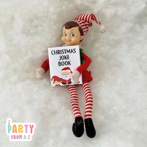 Op de afbeelding: Een rood-wit gestreepte elfpop die een klein boekje vasthoudt met de tekst "Christmas Joke Book" met een cartoon van Sinterklaas, elf en kind op de omslag.