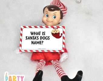Può includere: Un pupazzo di elfo a strisce rosse e bianche che tiene un biglietto bianco con il testo "Come si chiamano i cani di Babbo Natale?" L'elfo è sdraiato su un letto di neve bianca.