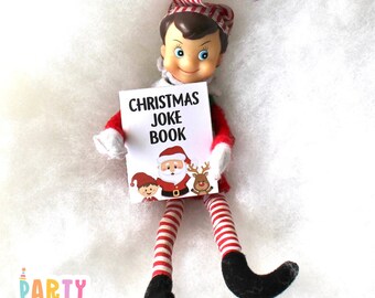 Può includere: Un pupazzo di elfo di Natale che tiene un libro intitolato "Christmas Joke Book". Il libro presenta un cartone animato di Babbo Natale, un elfo e una renna.