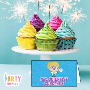 May include: A collection of six cupcakes with colorful frosting, including yellow, purple, pink, green, and blue, arranged on a white cake stand. Sparklers are lit above the cupcakes. A sign reads "Mad Scientist Cupcakes."