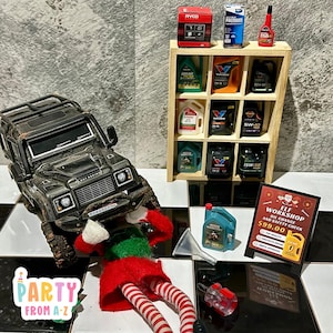 May include: A toy elf mechanic is working on a black toy Land Rover. The elf is wearing a red and green Christmas elf outfit. A wooden shelf with various toy oil bottles and containers is in the background. A sign reads "ELF WORKSHOP OIL CHANGE AND SAFETY CHECK £99.00".