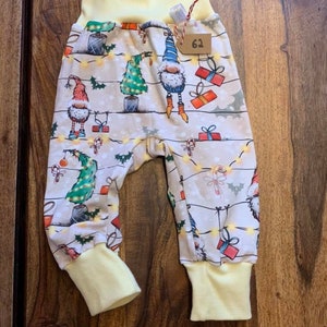 Könnte beinhalten: Gelbe und weiße Babyhose mit einem Weihnachts-Gnom-Druck. Die Hose hat einen gelben elastischen Bund und Bündchen. Der Druck zeigt Gnome, Weihnachtsbäume, Geschenke und Zuckerstangen. Die Hose hat ein Etikett mit der Aufschrift "62".