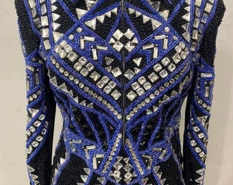 May include: Black equestrian show jacket with intricate blue and silver embellishments. The jacket features a zip-up front, a collar, and is adorned with a geometric pattern of rhinestones and beads. The number "12" is visible on the collar.