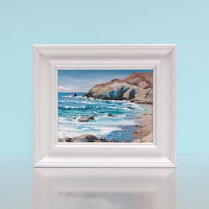 May include: A framed painting of a coastal scene. The artwork shows a rocky shoreline with turquoise water and a light blue sky. The white frame surrounds the painting, which is set against a light blue backdrop.