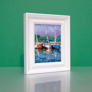 Könnte beinhalten: Gerahmtes Ölgemälde von Segelbooten in einem Hafen. Das Kunstwerk hat einen weißen Rahmen und zeigt Boote mit weißen Masten und farbigen Rümpfen, die sich im Wasser spiegeln. Der Hintergrund zeigt einen grünen Hügel und einen violetten Himmel.