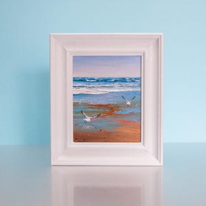 May include: A framed oil painting of a beach scene with two seagulls in flight. The artwork features a sandy shore, blue ocean waves, and a light blue sky. The painting is set in a white wooden frame.