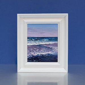 May include: A framed oil painting of a seascape. The artwork depicts ocean waves in shades of blue and white, under a sky of pink and blue hues. The painting is encased in a white rectangular frame, set against a blue background.