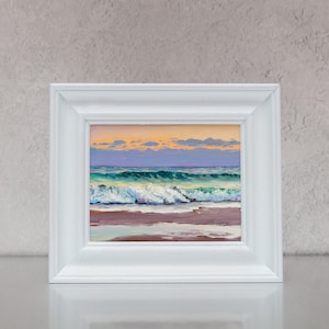 May include: A framed oil painting of a seascape. The artwork shows ocean waves in shades of turquoise and white, crashing onto a sandy shore. The sky transitions from orange to purple, with soft clouds. The frame is white.