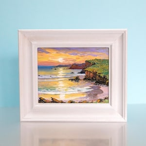 May include: A framed oil painting depicts a coastal scene at sunset. The artwork features a vibrant sky with orange and yellow hues, reflecting on the water. Green cliffs and a sandy beach are visible. The white frame complements the painting's colors.
