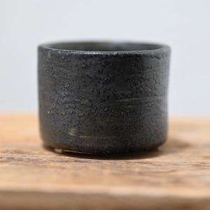 May include: A small, cylindrical ceramic cup with a dark charcoal black finish. The exterior surface features a rough, textured, and speckled appearance with subtle horizontal ridges circling the body of the vessel, creating a tactile look.