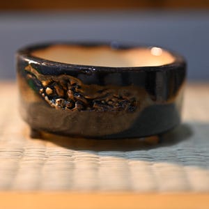 May include: A small, round ceramic bonsai pot with a glossy, dark navy blue and brown glaze. The exterior features a textured, rugged relief pattern along the side. The interior is a smooth, light cream color, and the pot stands on small feet.