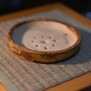 May include: A round, shallow ceramic dish with a textured, mottled brown exterior and a light beige interior. The base of the dish features a circular pattern of small, evenly spaced drainage holes, suggesting its use as a plant pot saucer.
