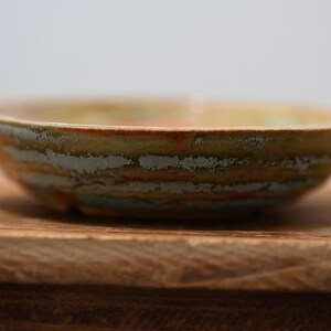 May include: A shallow, circular ceramic bowl featuring a textured, multi-colored glaze with horizontal bands of muted green, earthy brown, and hints of orange. The rim is slightly uneven, and the exterior surface displays a rustic, layered finish.