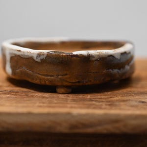 May include: A small, shallow, circular ceramic bowl with a textured, earthy brown exterior and a thick, uneven white glaze dripping over the top rim. The bowl features a low-profile design with small, integrated feet at the base for stability.