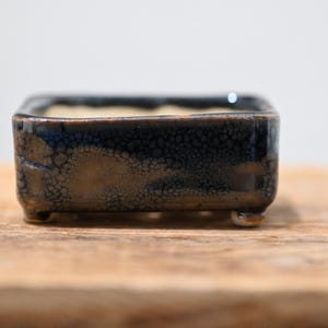 Può includere: Un vaso rettangolare in ceramica per bonsai con smalto reattivo blu scuro e marrone caratterizzato da una visibile texture craquelé. Il contenitore presenta un bordo liscio leggermente arrotondato ed è sostenuto da quattro piedini corti.