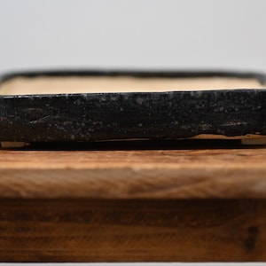 May include: A rectangular bonsai pot with a textured, matte black exterior finish and a smooth, light beige interior. The low-profile container features slightly raised feet at the base, providing a stable foundation for plant cultivation.