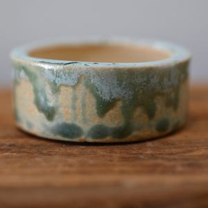 May include: A small, round ceramic bowl with a low profile and an open top. The exterior features a reactive glaze with a blend of earthy tan, sage green, and muted blue tones that drip down the sides, creating a textured, multi-colored finish.