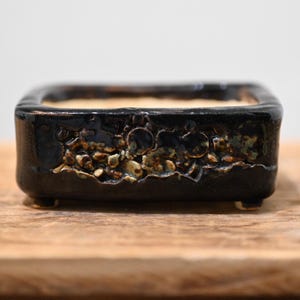 May include: A rectangular ceramic bonsai pot with a glossy black glaze. The exterior features a textured, cratered band of mottled brown and tan glaze across the center. The pot is supported by four small, short feet at the base.