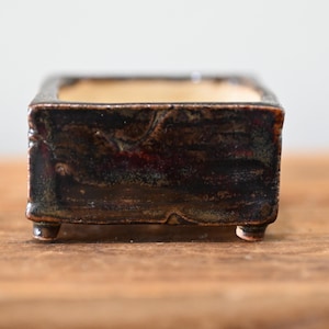 May include: A small, square ceramic planter with a dark, mottled brown and black glazed exterior. The vessel features a smooth, light-colored interior and is supported by four short, stubby feet at the base, showcasing a textured, rustic finish.