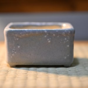 May include: A rectangular ceramic planter with a light blue, speckled glaze finish. The vessel features rounded corners, a smooth rim, and four small, short feet at the base, providing a stable foundation for small plants or decorative items.