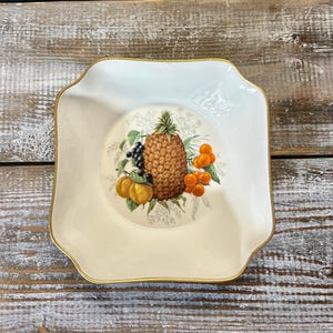 May include: A white square ceramic dish with a gold rim. The dish features a painted design of a pineapple, cherries, and other fruit.