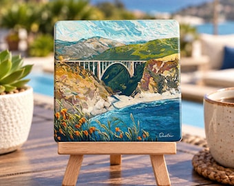 May include: A square art print on a wooden easel featuring a painting of the Bixby Creek Bridge in Big Sur, California. The scene depicts a white arched bridge spanning a canyon above a blue ocean, with orange poppies in the foreground.