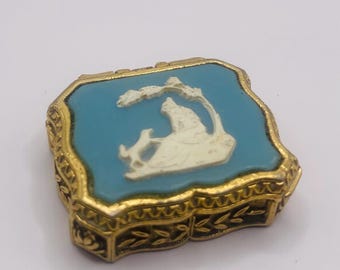 May include: A small, square, gold-coloured trinket box with a blue enamel top and a white cameo of a woman and a dog. The box has a decorative, ornate border.