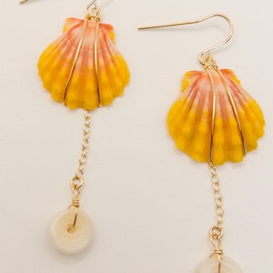Puede incluir: Un par de pendientes de oro con una concha de mar amarilla y naranja con un diseño de alambre dorado y un dije de botón blanco que cuelga de una cadena de oro.