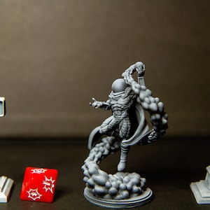 Puede incluir: Una figura en miniatura de pl&aacute;stico gris de un superh&eacute;roe con una m&aacute;scara negra y una capa, de pie sobre una base de pl&aacute;stico gris con un efecto de humo arremolinado. La figura est&aacute; en una pose de acci&oacute;n din&aacute;mica. Hay una farola de pl&aacute;stico gris y un sem&aacute;foro de pl&aacute;stico gris a cada lado de la figura. Un dado rojo con puntos blancos est&aacute; delante de la figura.
