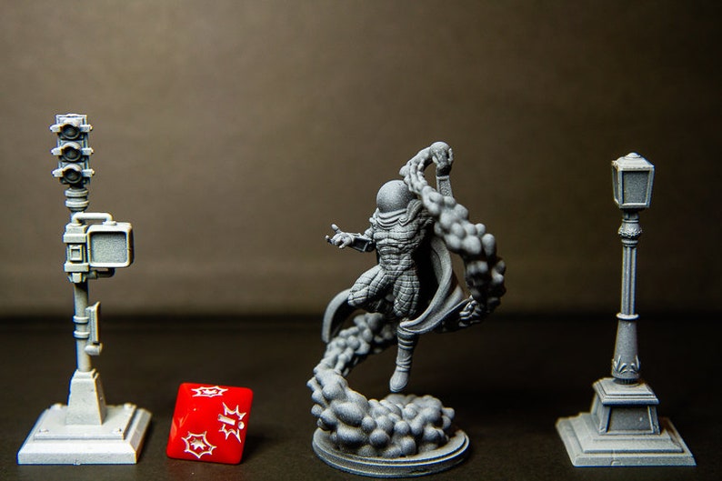 Puede incluir: Una figura en miniatura de pl&aacute;stico gris de un superh&eacute;roe con una m&aacute;scara negra y una capa, de pie sobre una base de pl&aacute;stico gris con un efecto de humo arremolinado. La figura est&aacute; en una pose de acci&oacute;n din&aacute;mica. Hay una farola de pl&aacute;stico gris y un sem&aacute;foro de pl&aacute;stico gris a cada lado de la figura. Un dado rojo con puntos blancos est&aacute; delante de la figura.