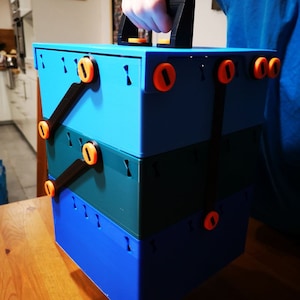 May include: A stackable, blue, green and black plastic storage container with a black handle. The container has orange buttons on the sides.