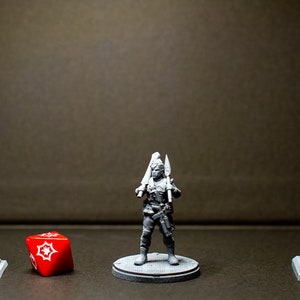 Può includere: Una figura in miniatura grigia di una persona che tiene due spade, in piedi su una base grigia. La figura è davanti a un lampione grigio a sinistra e un semaforo grigio a destra. Un dado rosso con segni bianchi è in primo piano.