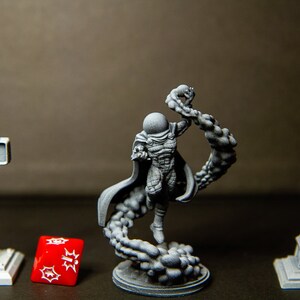 Puede incluir: Una miniatura gris de un personaje de superh&eacute;roe con una capa y un dise&ntilde;o de telara&ntilde;a, impresa en 3D. El personaje est&aacute; de pie sobre una base con un dise&ntilde;o de nube. Hay dos farolas grises impresas en 3D y un dado rojo sobre la mesa.