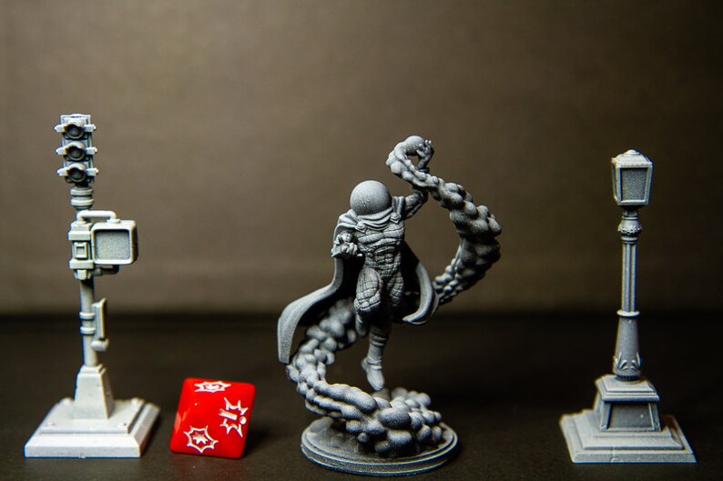 Puede incluir: Una miniatura gris de un personaje de superh&eacute;roe con una capa y un dise&ntilde;o de telara&ntilde;a, impresa en 3D. El personaje est&aacute; de pie sobre una base con un dise&ntilde;o de nube. Hay dos farolas grises impresas en 3D y un dado rojo sobre la mesa.