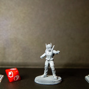 Op de afbeelding: Een grijs miniatuurfiguur van een persoon met een helm en een wapen staat op een grijze basis. De figuur is omringd door drie grijze miniatuurstructuren, waaronder een lantaarnpaal, een verkeerslicht en een gebouw. Een rode dobbelsteen met witte symbolen bevindt zich op de voorgrond.