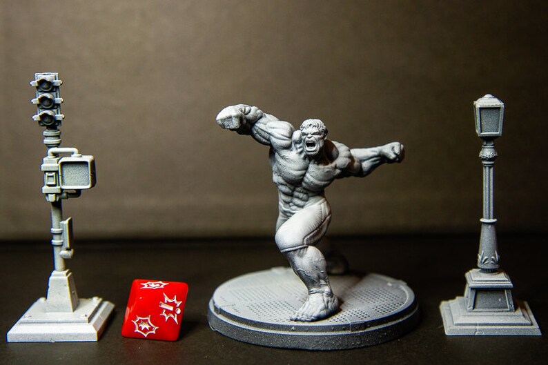 May include: A gray plastic miniature figurine of the Hulk in a fighting pose. The Hulk is standing on a gray plastic base. There is a gray plastic streetlight and a gray plastic traffic light on either side of the Hulk. A red plastic dice with white spots is in front of the Hulk.