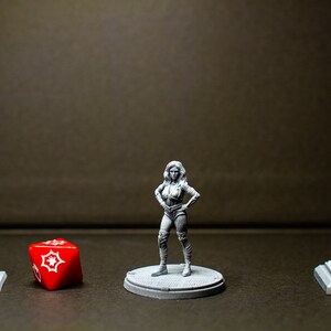 Puede incluir: Una figura en miniatura gris de una mujer de pie sobre una base gris. La figura lleva un atuendo negro y gris. Un dado rojo con estrellas blancas está en primer plano. Dos farolas en miniatura grises están a cada lado de la figura.