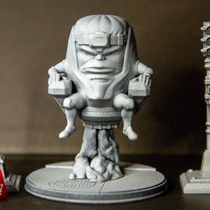 Può includere: Una miniatura grigia stampata in 3D di un personaggio seduto su un trono, con un lampione e un semaforo su entrambi i lati. Il personaggio ha una testa grande e un corpo piccolo, e indossa un casco. Il lampione e il semaforo sono entrambi su basi.