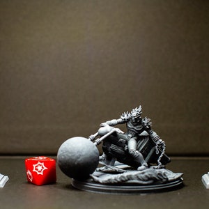 Peut inclure: Une miniature en 3D imprimée en gris d'une figurine à moto avec une grosse boule attachée à l'avant. La figurine porte un casque et une armure. La miniature est sur une base grise. Il y a un lampadaire gris et un feu de circulation gris de chaque côté de la miniature. Un dé rouge à 20 faces se trouve devant la miniature.