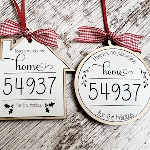 May include: Two wooden Christmas ornaments, one shaped like a house and the other round. Both ornaments are white with black lettering that reads "There's no place like home 54937 for the holidays". The ornaments are decorated with red and white gingham ribbon.