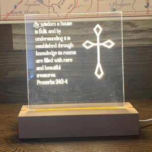 May include: A white illuminated acrylic sign with a cross design and a quote from Proverbs 24:3-4: "By wisdom a house is built and by understanding it is established through knowledge its rooms are filled with rare and beautiful treasures. Proverbs 24:3-4"