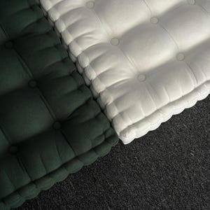 “Top-down view of two thick tufted floor cushions placed side by side on a dark carpet, one in deep green and the other in off-white, with soft fabric texture and evenly spaced button details.