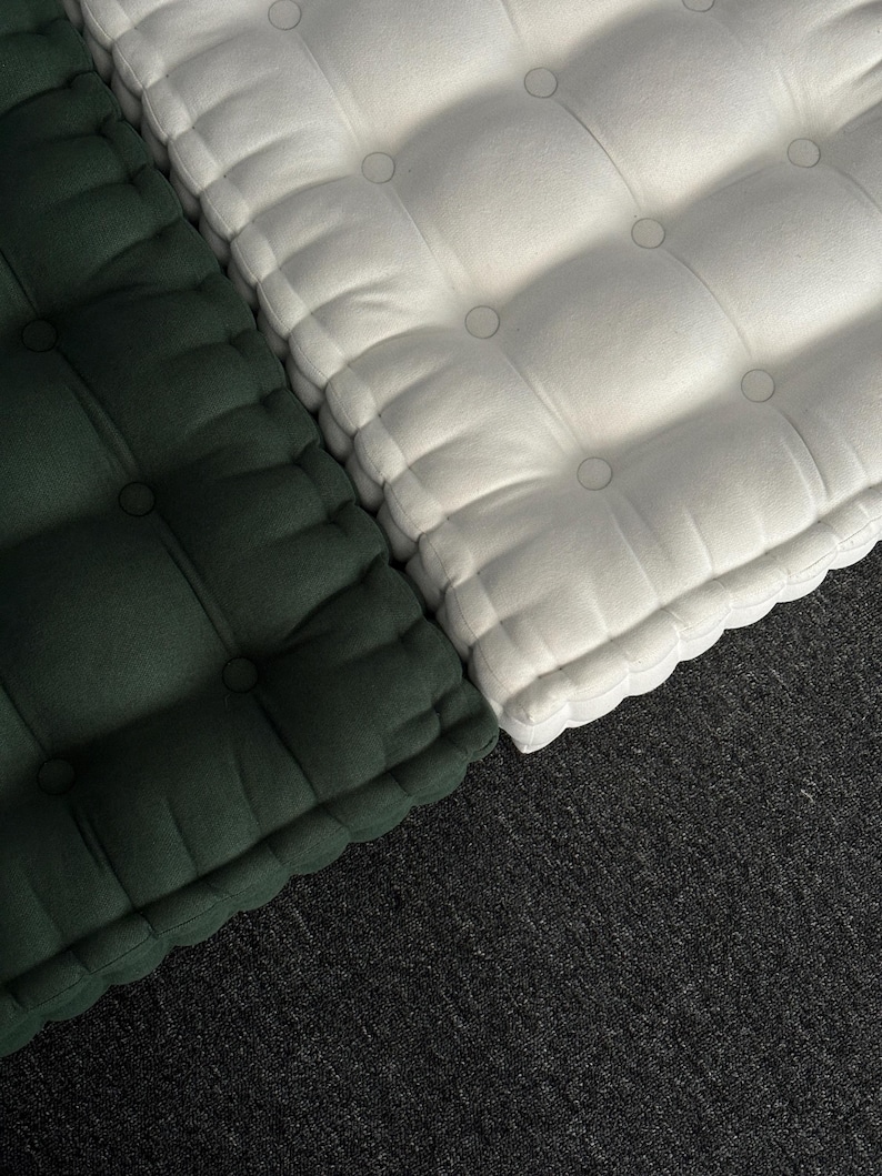 “Top-down view of two thick tufted floor cushions placed side by side on a dark carpet, one in deep green and the other in off-white, with soft fabric texture and evenly spaced button details.