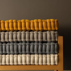 A wooden bench holding four neatly stacked, thick tufted cushions in warm mustard yellow, light gray, dark gray, and off-white, set against a smooth gray wall.