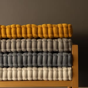 May include: A stack of four tufted cushions in shades of yellow, gray, and off-white. The cushions are rectangular and have a ribbed texture. They are stacked on a light-colored wooden frame, with a dark background.